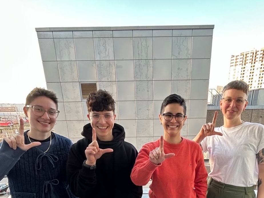 Vorstand LesbenRing e.V. 2025 Vier lächelnde Personen stehen nebeneinander im Freien vor einer hellen Gebäudewand; sie tragen Freizeitkleidung und schauen direkt in die Kamera.